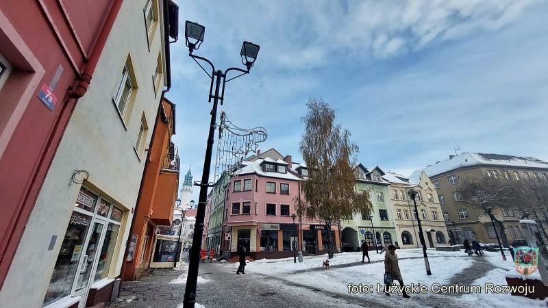 Widok na kamienice miejskie i zaśnieżony trakt. Na pierwszym planie nowa lampa, w tle przechodnie.